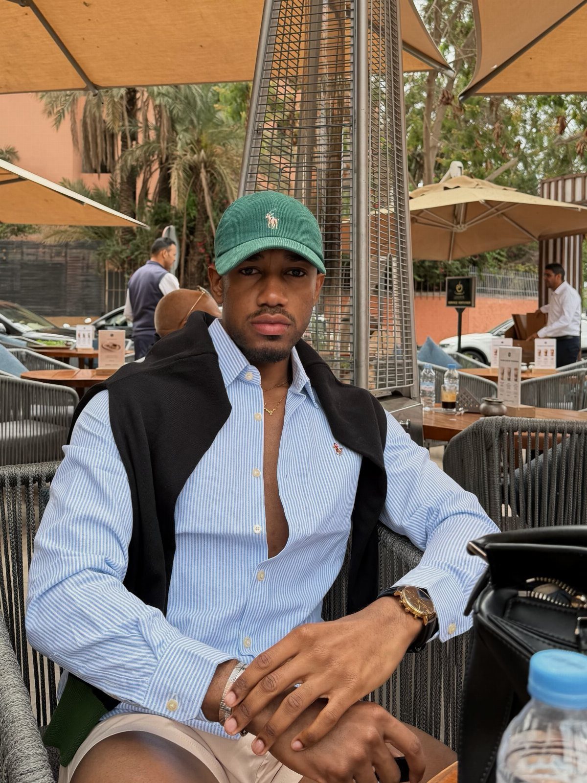 A man wearing a light blue striped shirt and a green cap sitting at an outdoor cafe.