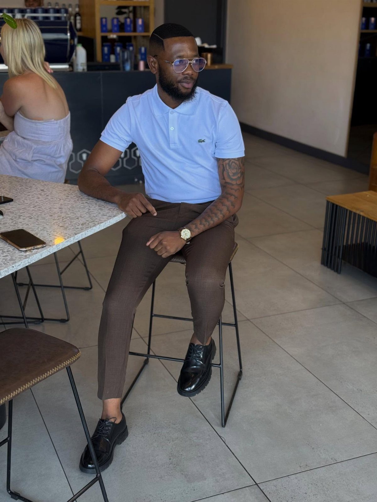 Man in a light blue polo shirt and brown trousers sitting on a stool in a cafe.