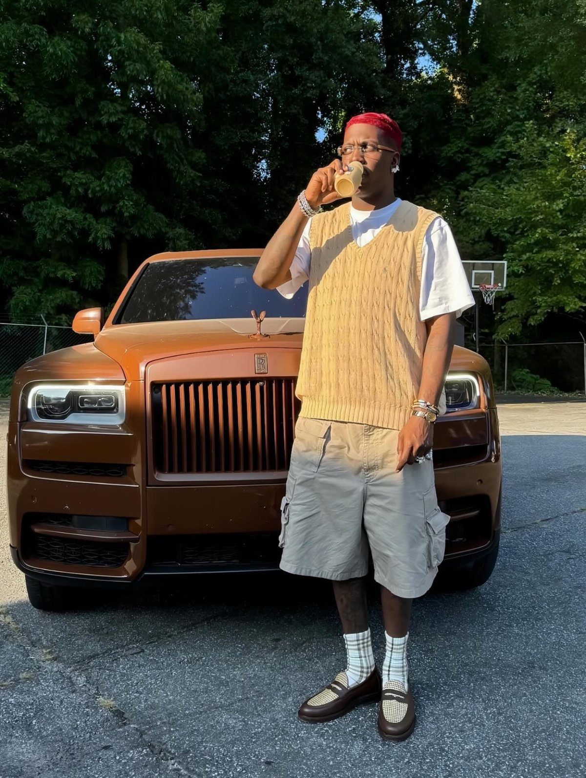 A man in a sweater vest and cargo shorts standing in front of a brown car.