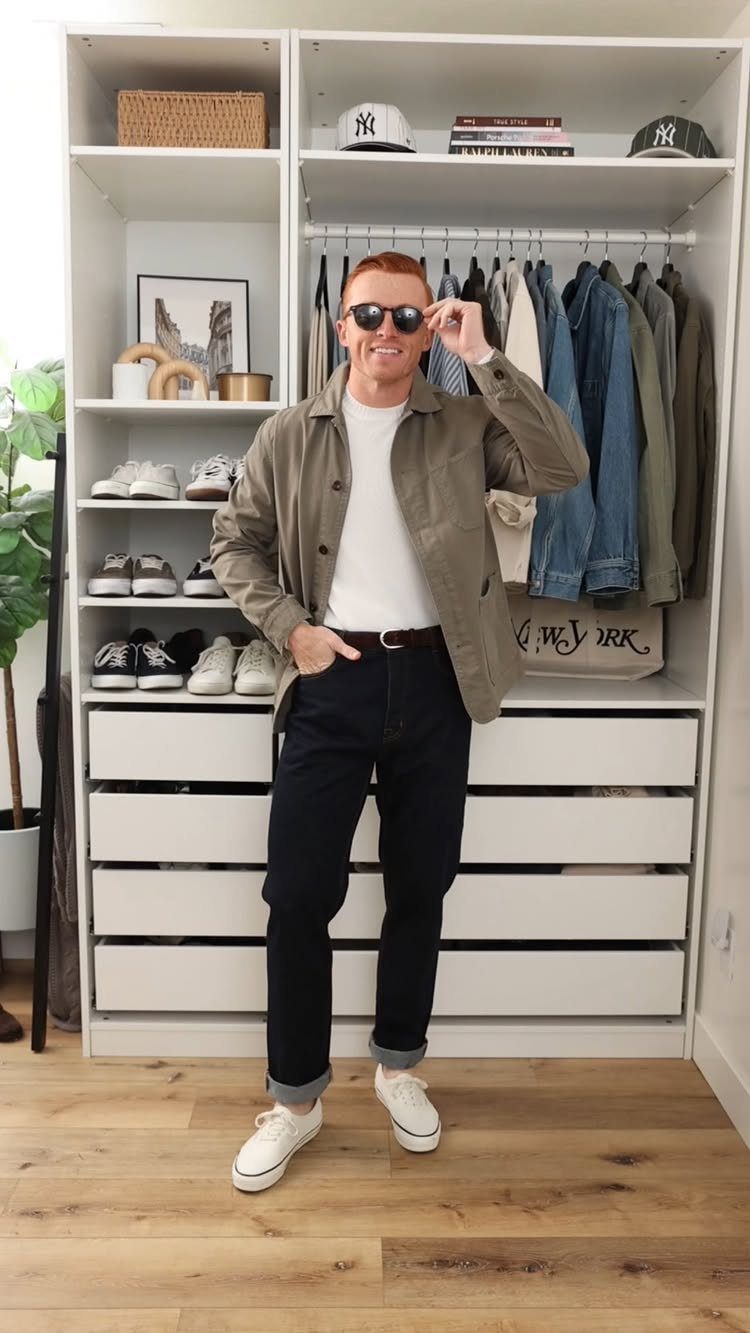 Man wearing a white t-shirt, olive overshirt, dark jeans, and white sneakers standing in a closet.