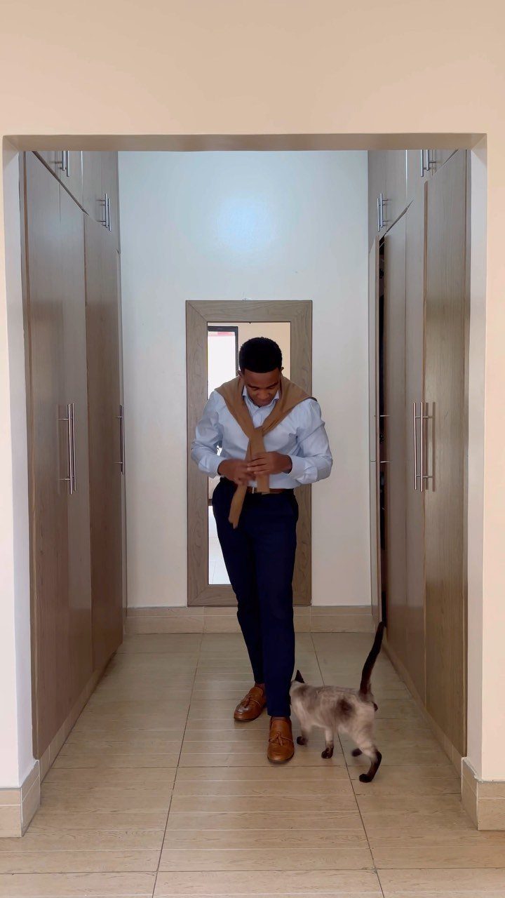 Man in light blue shirt and navy trousers standing in a hallway with a cat.