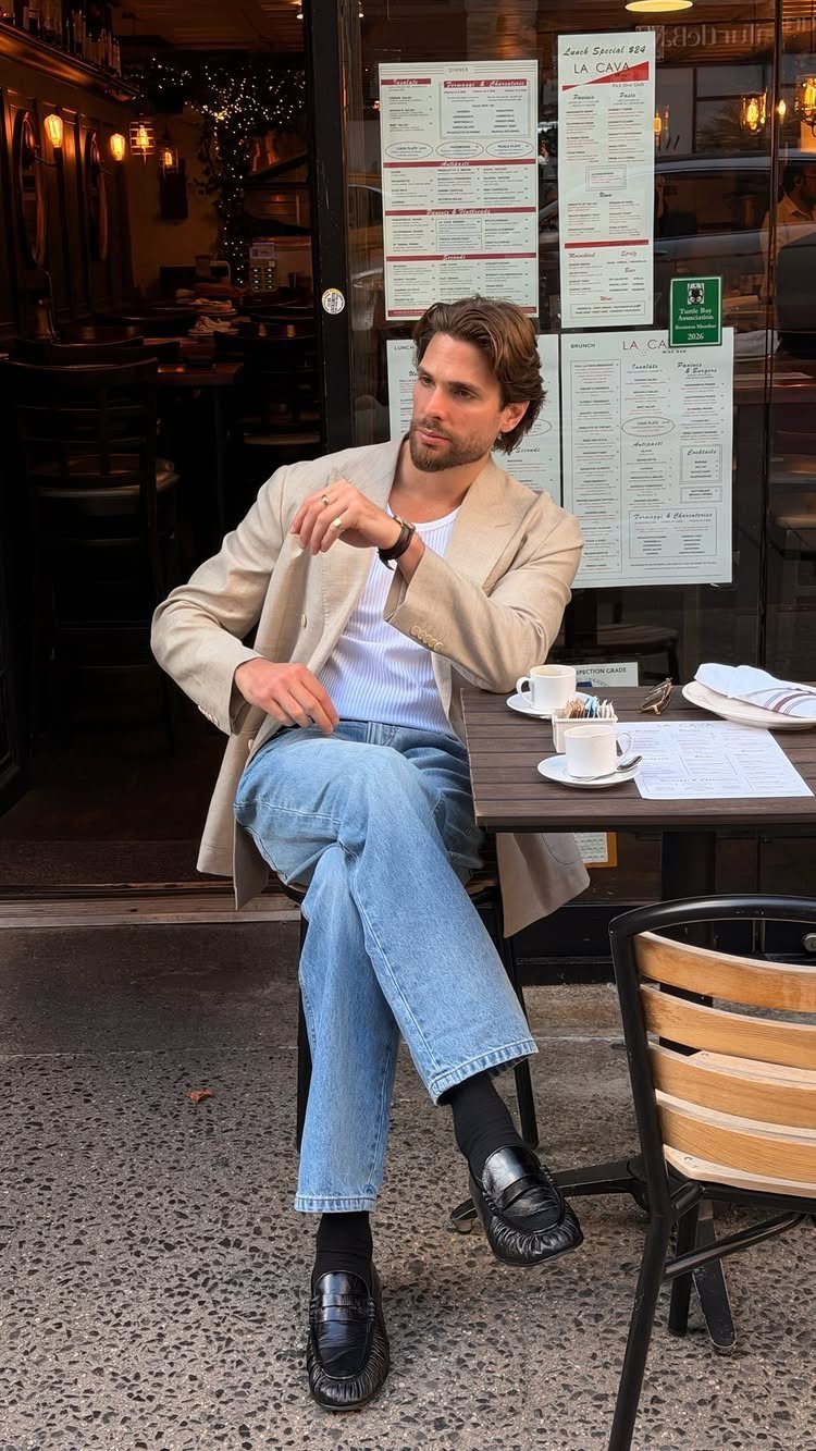 A man sitting at a cafe wearing a beige blazer, white t-shirt, and blue jeans.