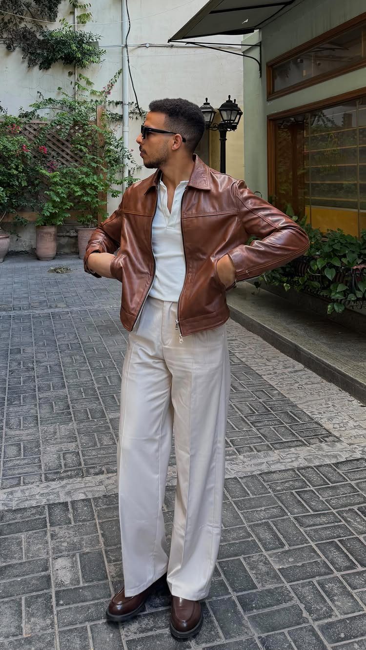 Man in a brown leather jacket, white henley, and cream trousers standing on a street.