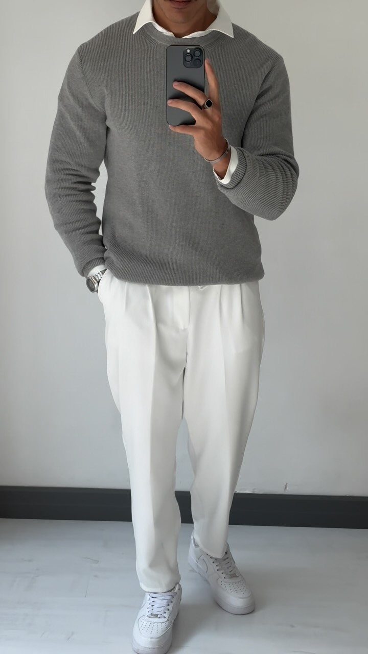 A person wearing a grey knit sweater, white collared shirt, and white trousers.
