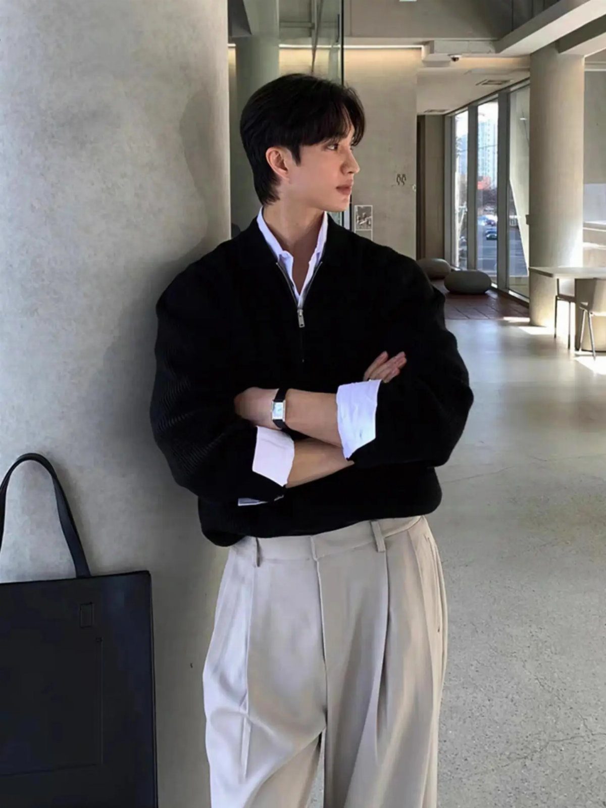 Man in a black knit sweater layered over a white shirt and beige trousers leaning against a pillar.