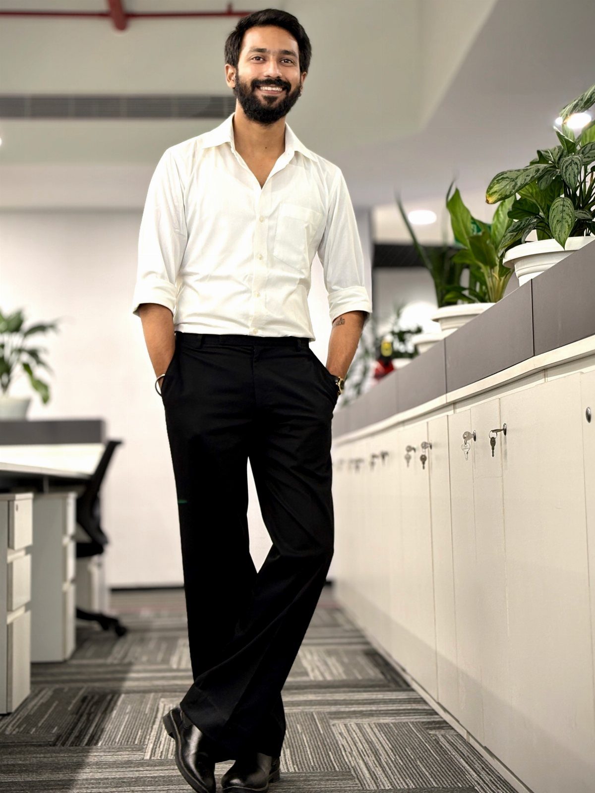 A smiling man in a white shirt and black trousers posing in a modern office.