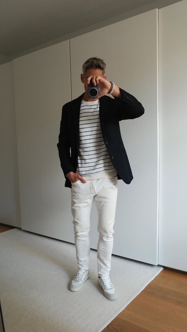 Man posing in a black blazer, striped shirt, white jeans, and white sneakers.