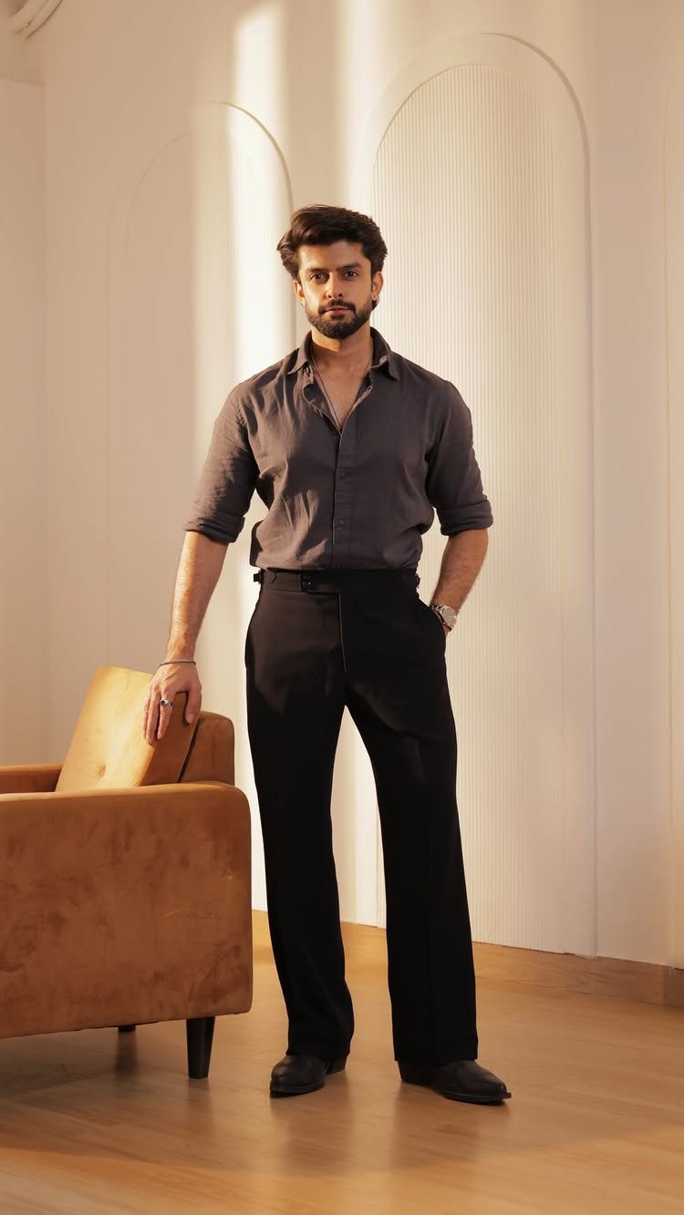 A man wearing a grey button-down shirt and black dress pants standing in a minimalist room.