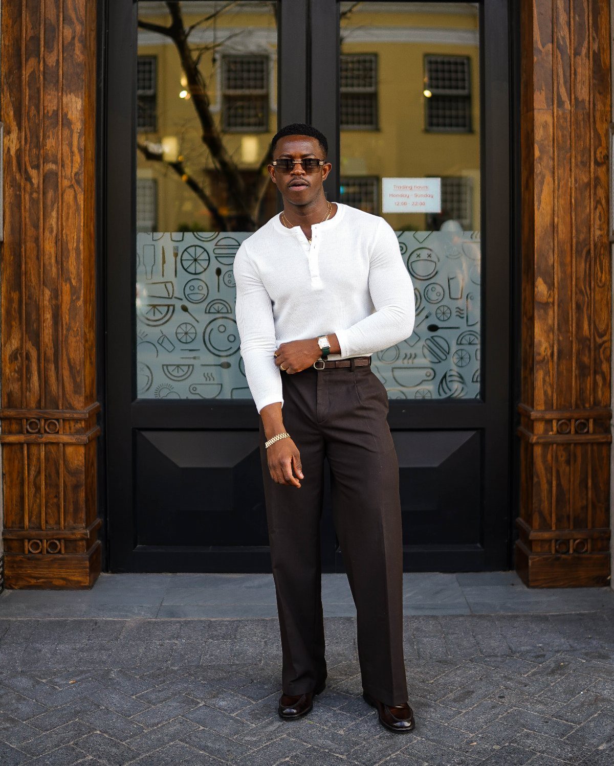 Man wearing a white henley shirt and dark brown trousers standing before dark doors.