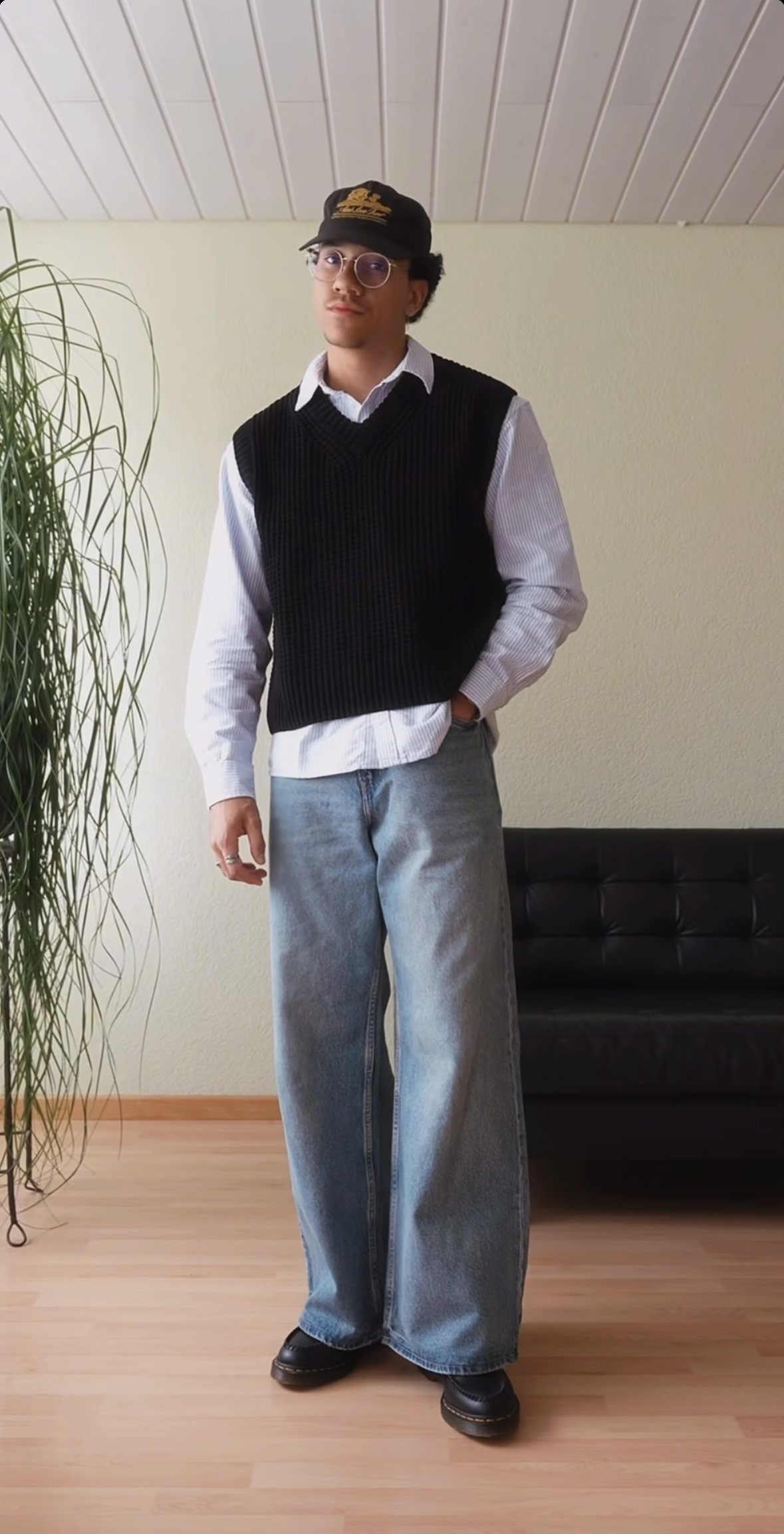 Man wearing a black sweater vest over a white long-sleeve shirt and wide denim jeans.