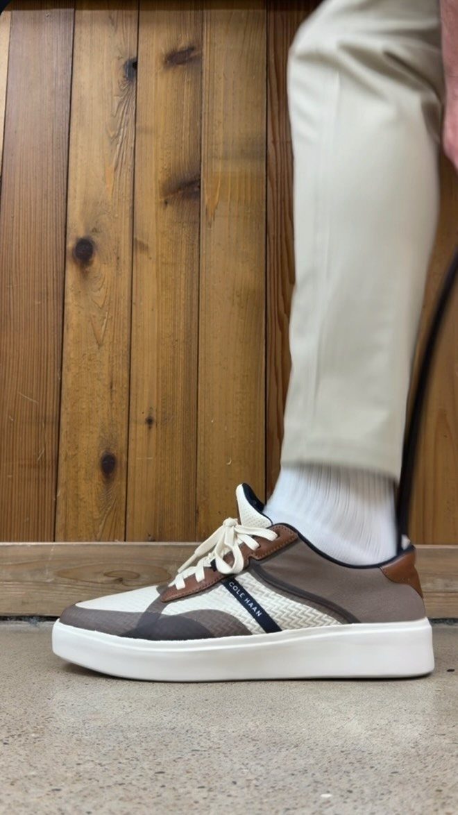 A person wearing beige trousers and brown and cream casual sneakers against a wooden wall.