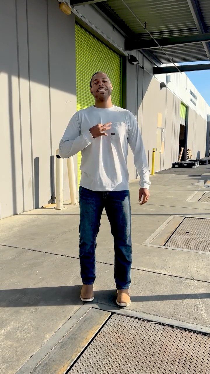 A man smiling while wearing a white long-sleeved shirt, blue jeans, and tan shoes outdoors.