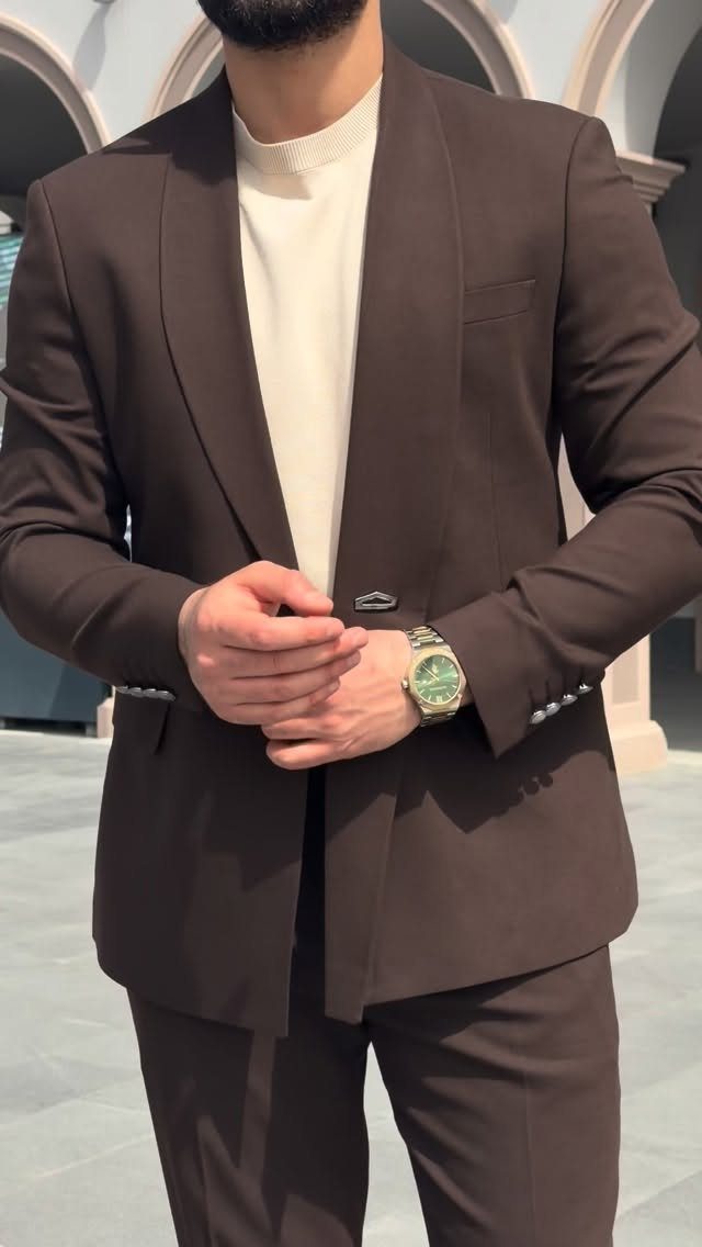 Man in a dark brown suit jacket and cream sweater standing outdoors.