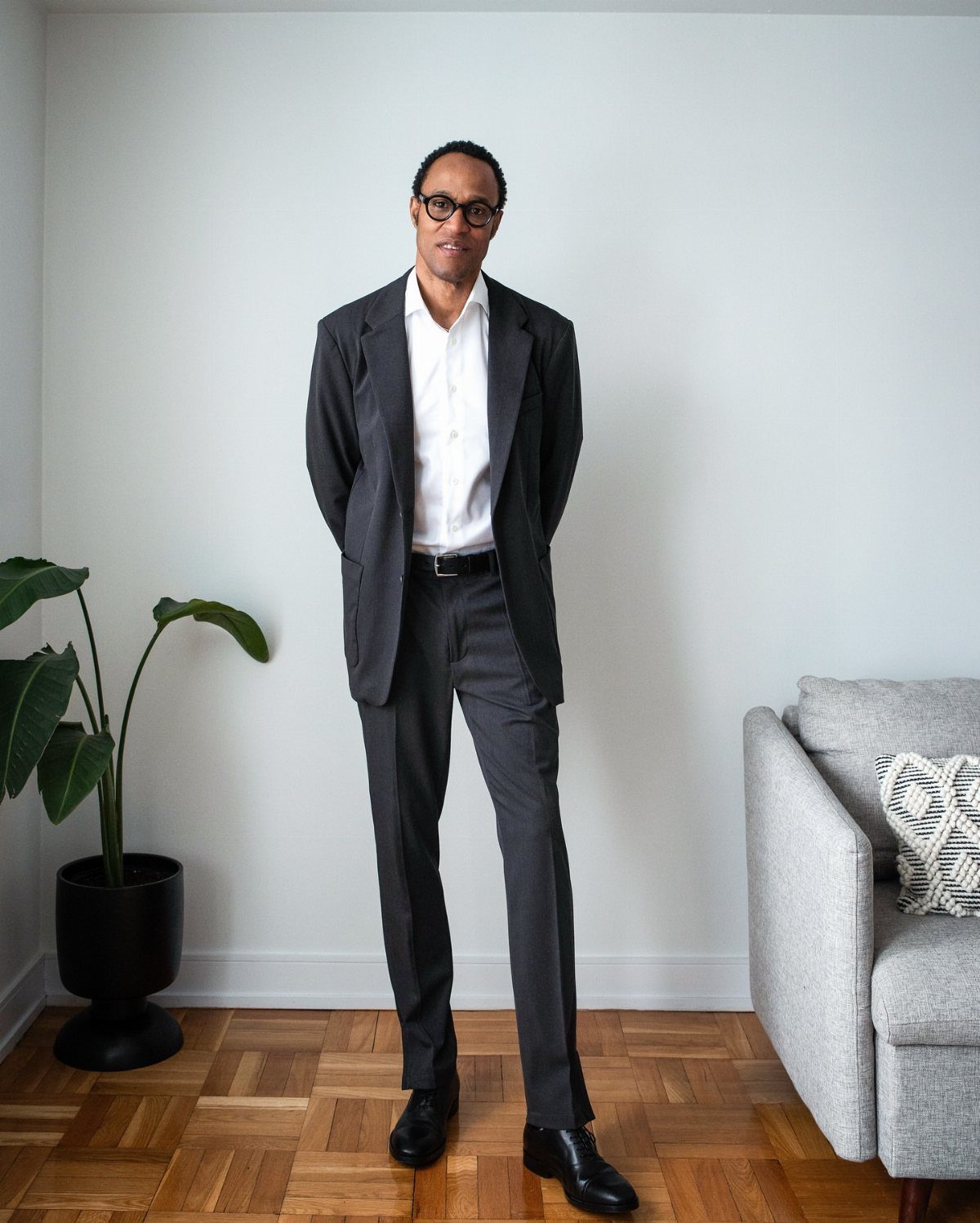 A man wearing a grey suit and white shirt stands in a bright room with a plant.