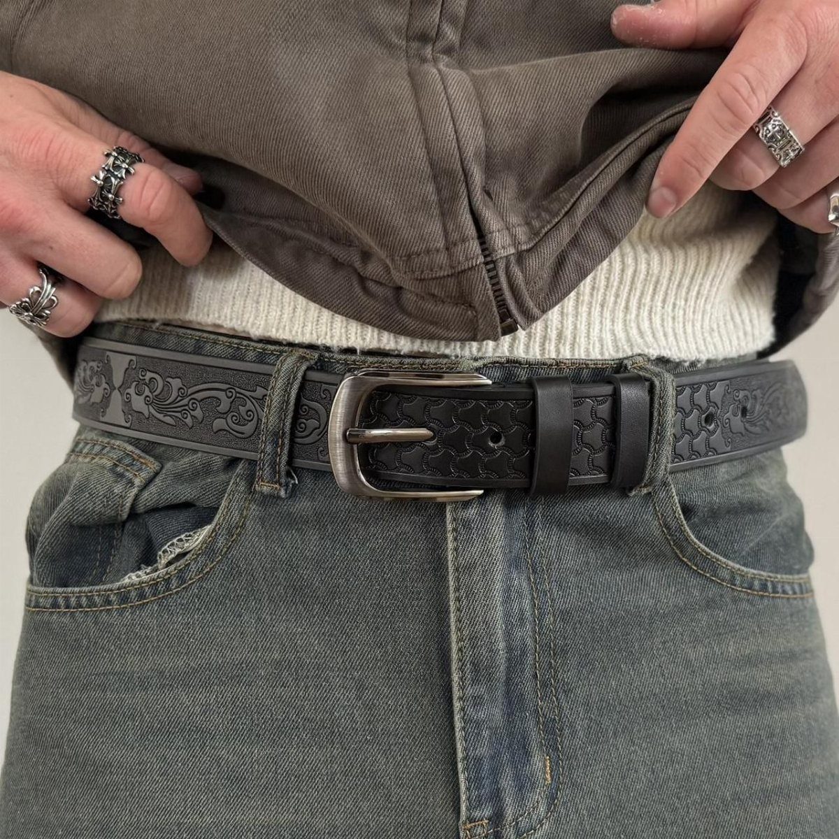 A person wearing blue denim jeans with a decorative black leather belt and silver rings.