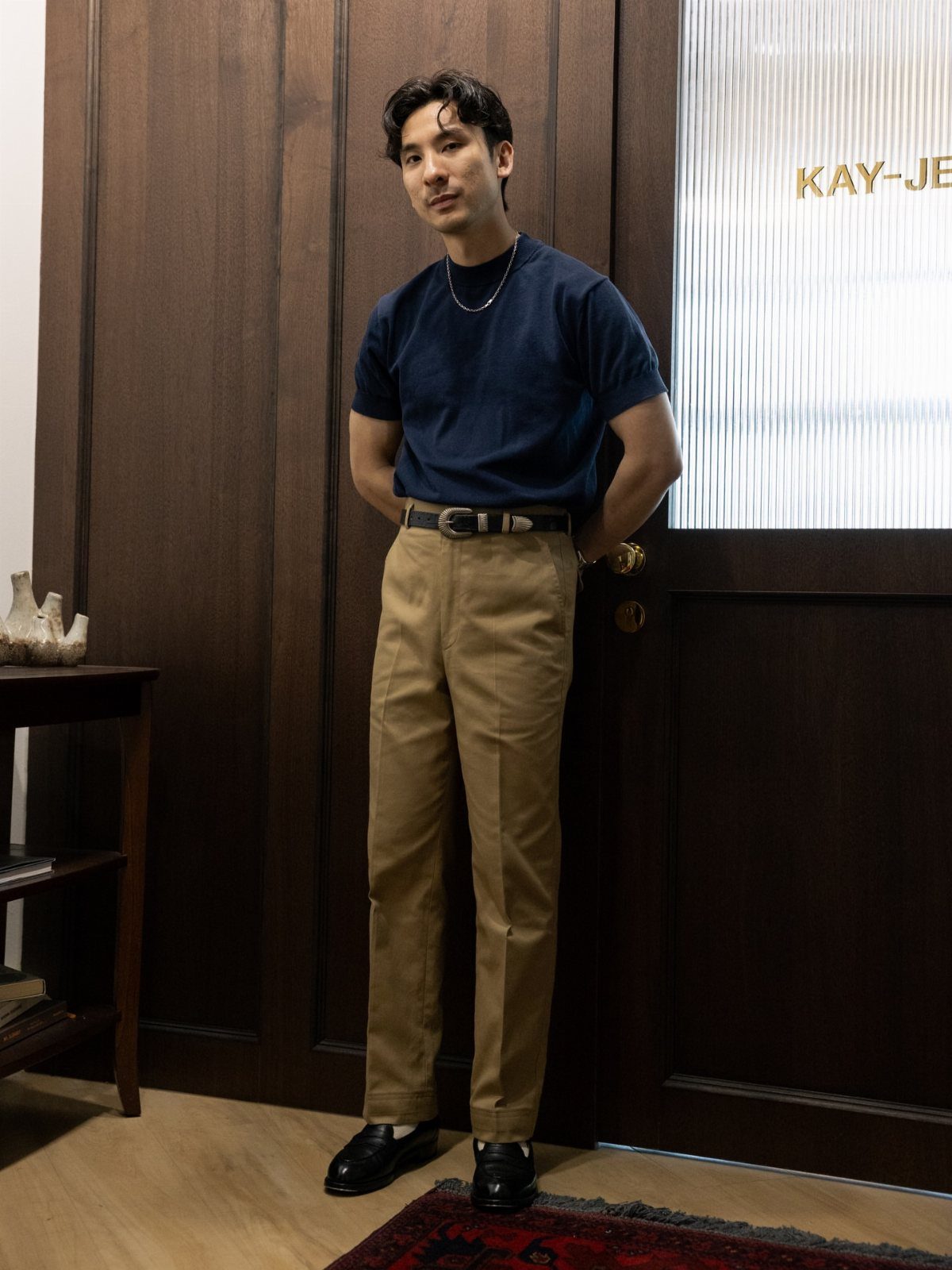 Man wearing a navy t-shirt and tan chino trousers standing in front of a wooden door.