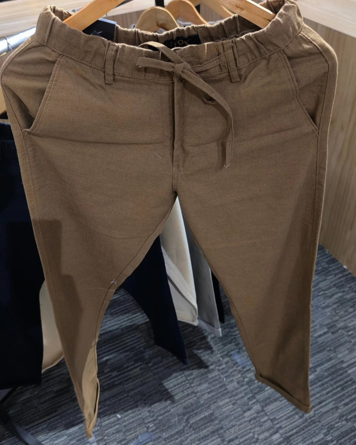 Tan drawstring trousers hanging on a wooden hanger in a retail store.