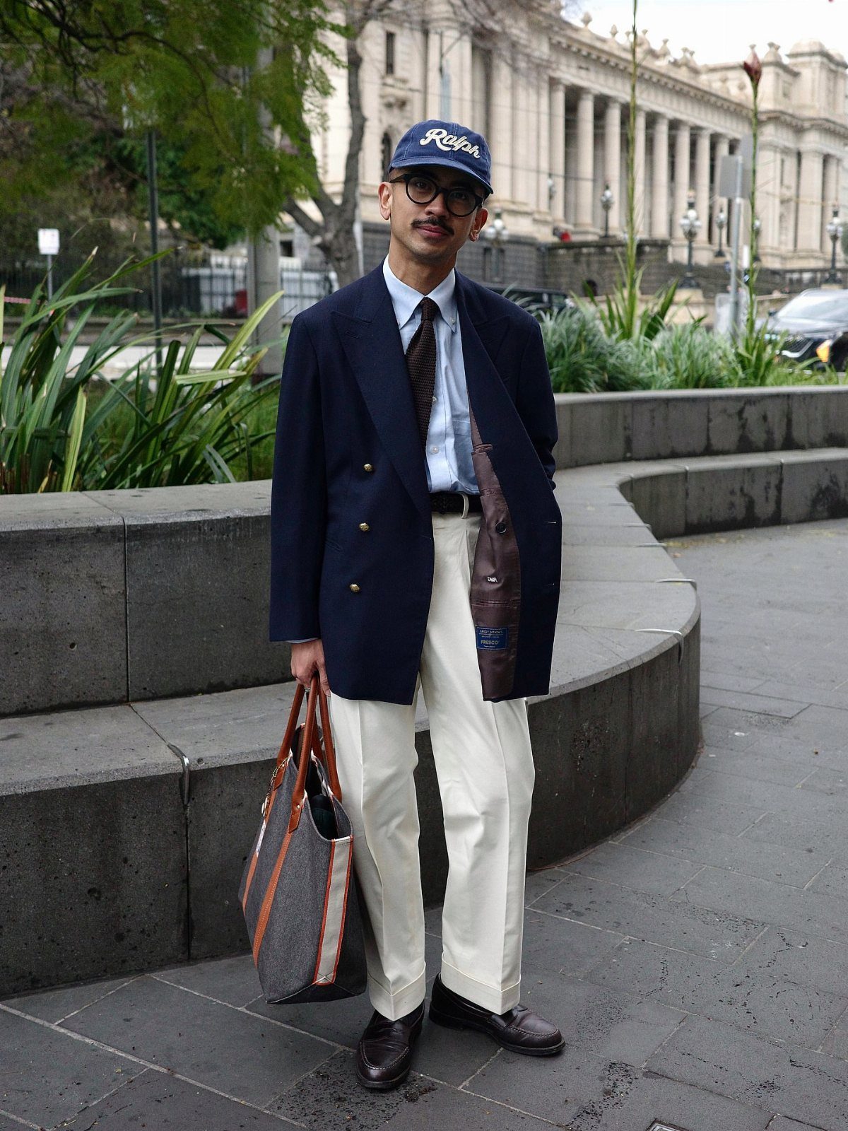 A man posing in a navy blue blazer, light blue shirt, tie, and cream trousers.