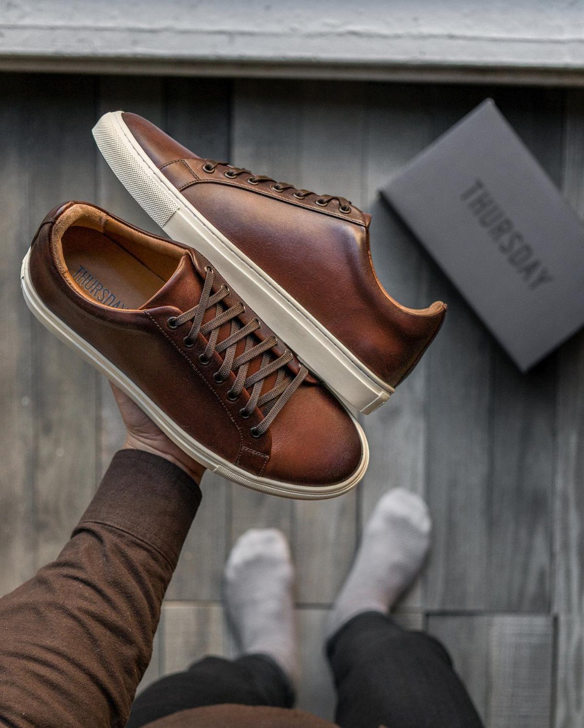 A person holding up two brown leather sneakers with white soles over a wooden floor.