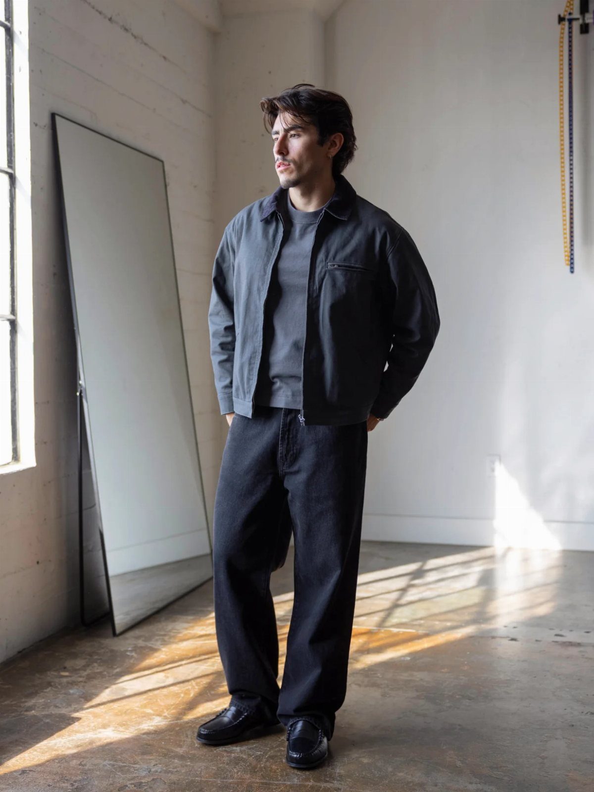 A man wearing a dark grey structured jacket, a grey t-shirt, and black trousers standing in a bright room.