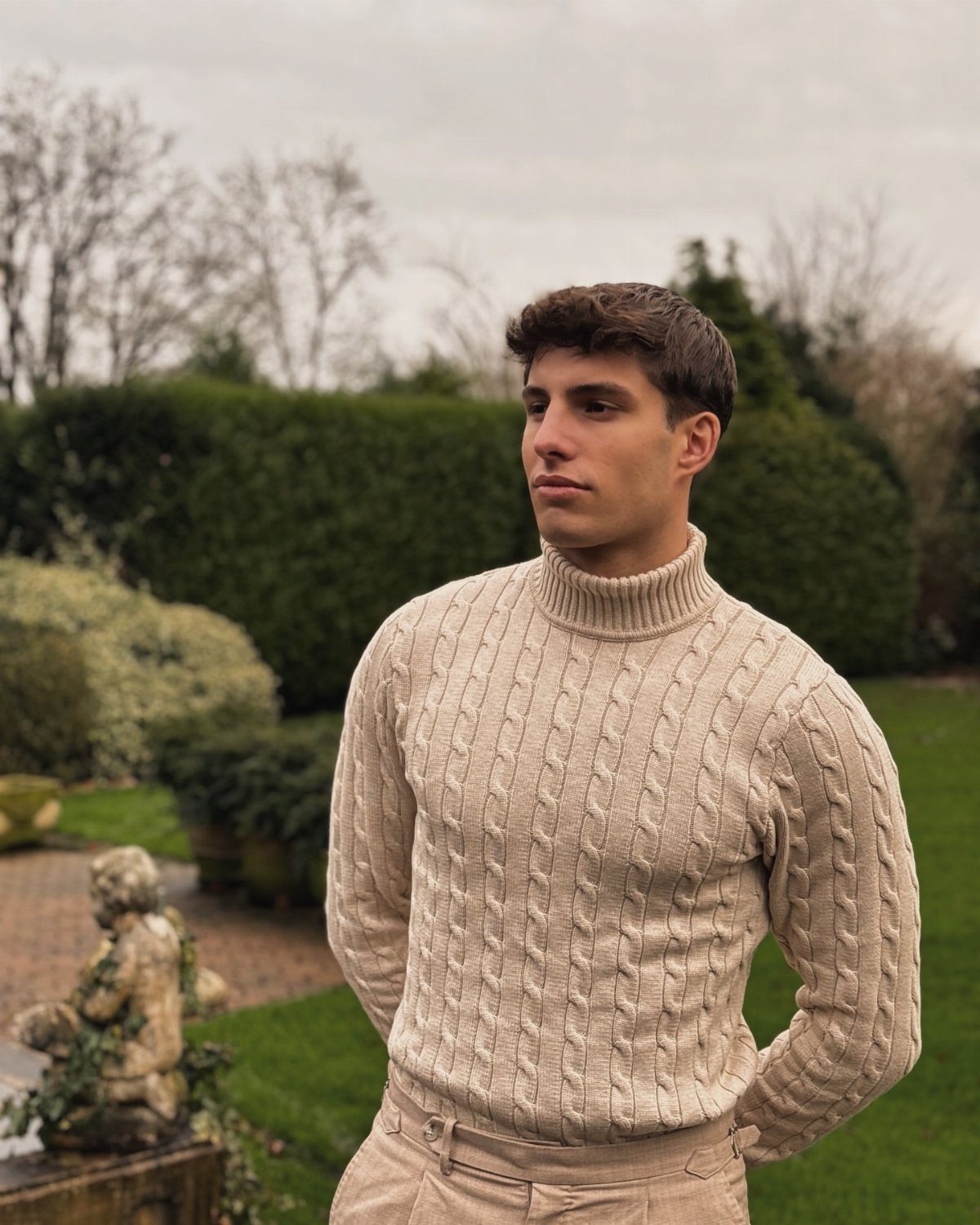 A man wearing a beige cable-knit turtleneck sweater standing in a lush green garden.