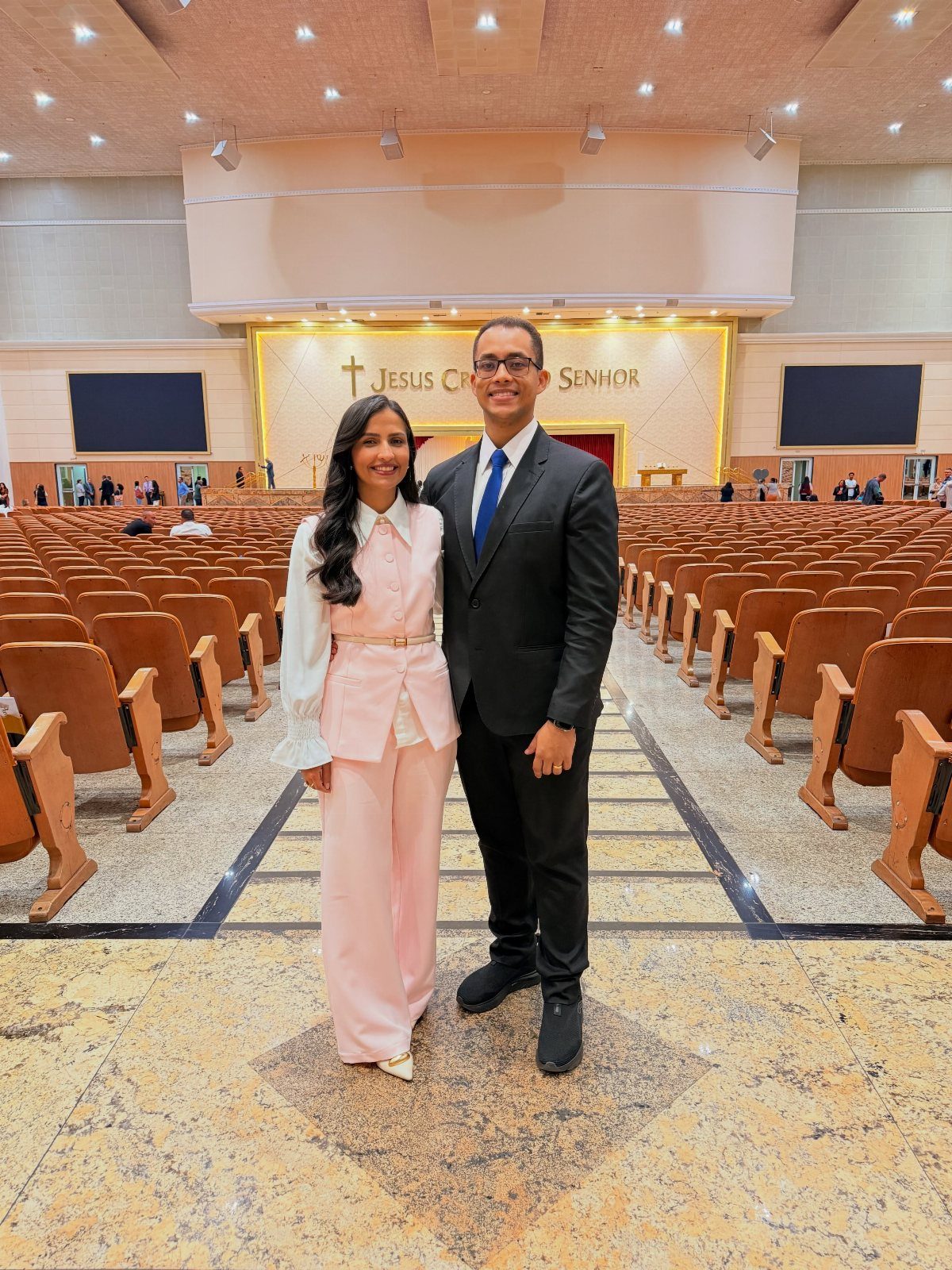 Man in a dark formal suit and tie standing with a woman in a pink outfit inside a church.