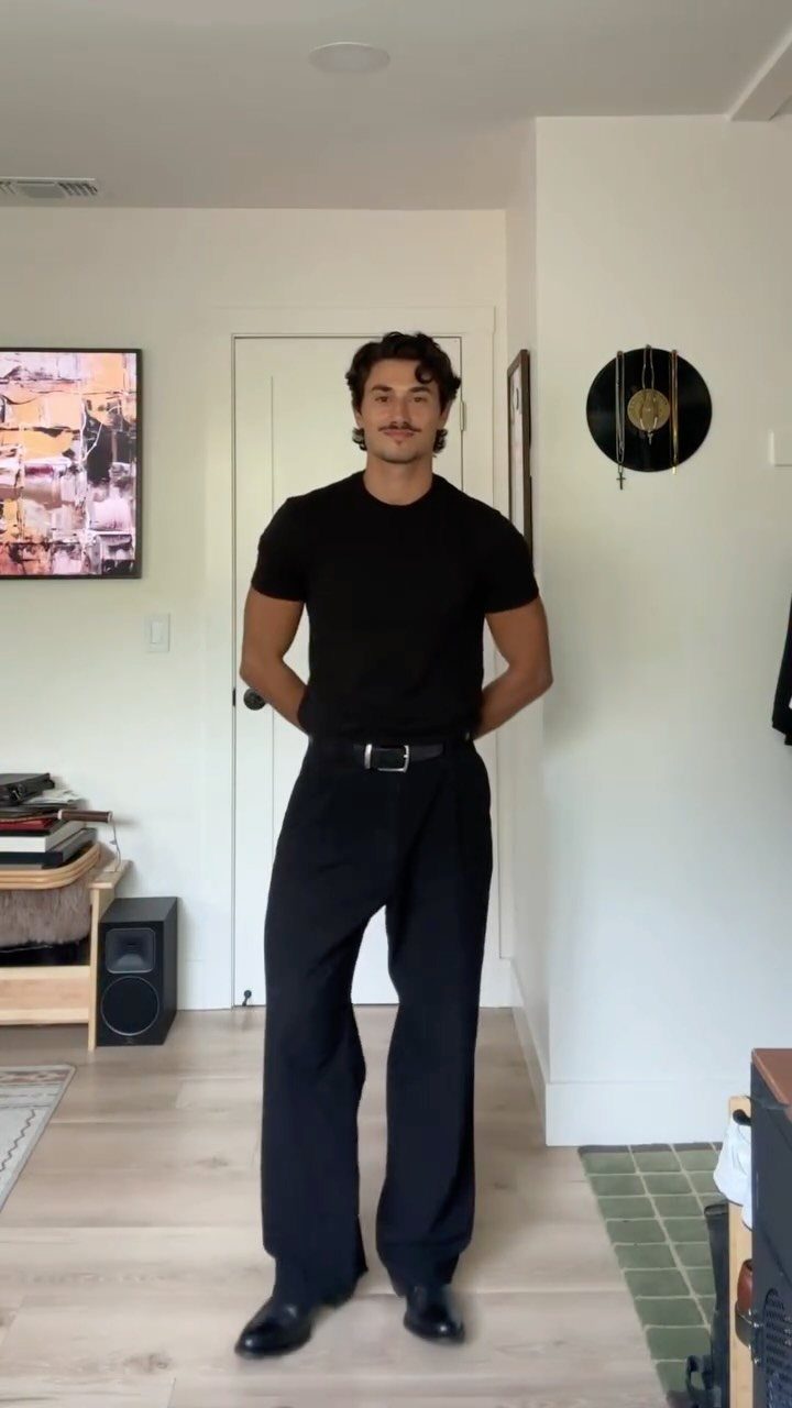 A man posing in a black t-shirt, wide-leg black trousers, and black dress shoes.