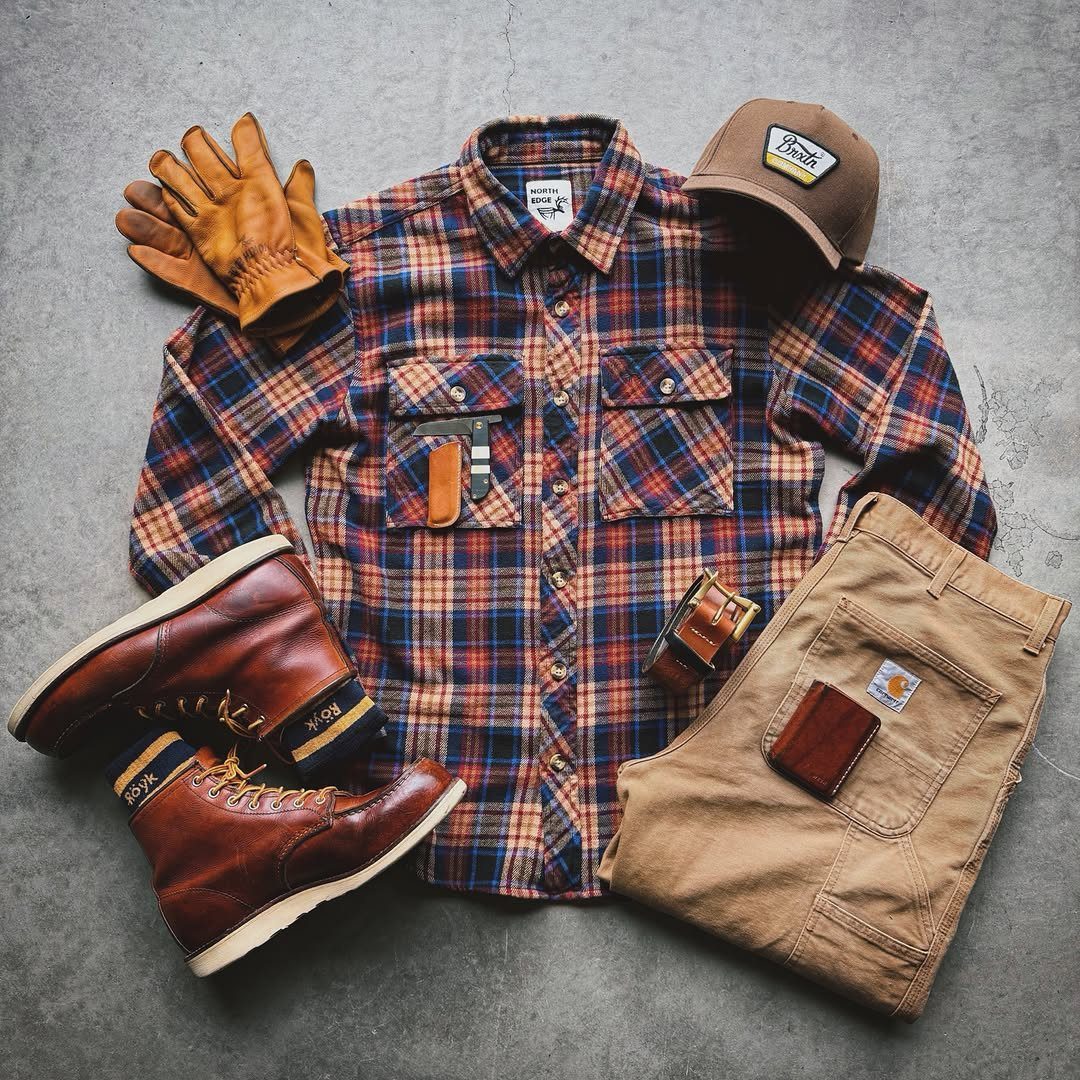 Plaid flannel shirt, tan work pants, leather boots, and work gloves arranged in a flat lay.