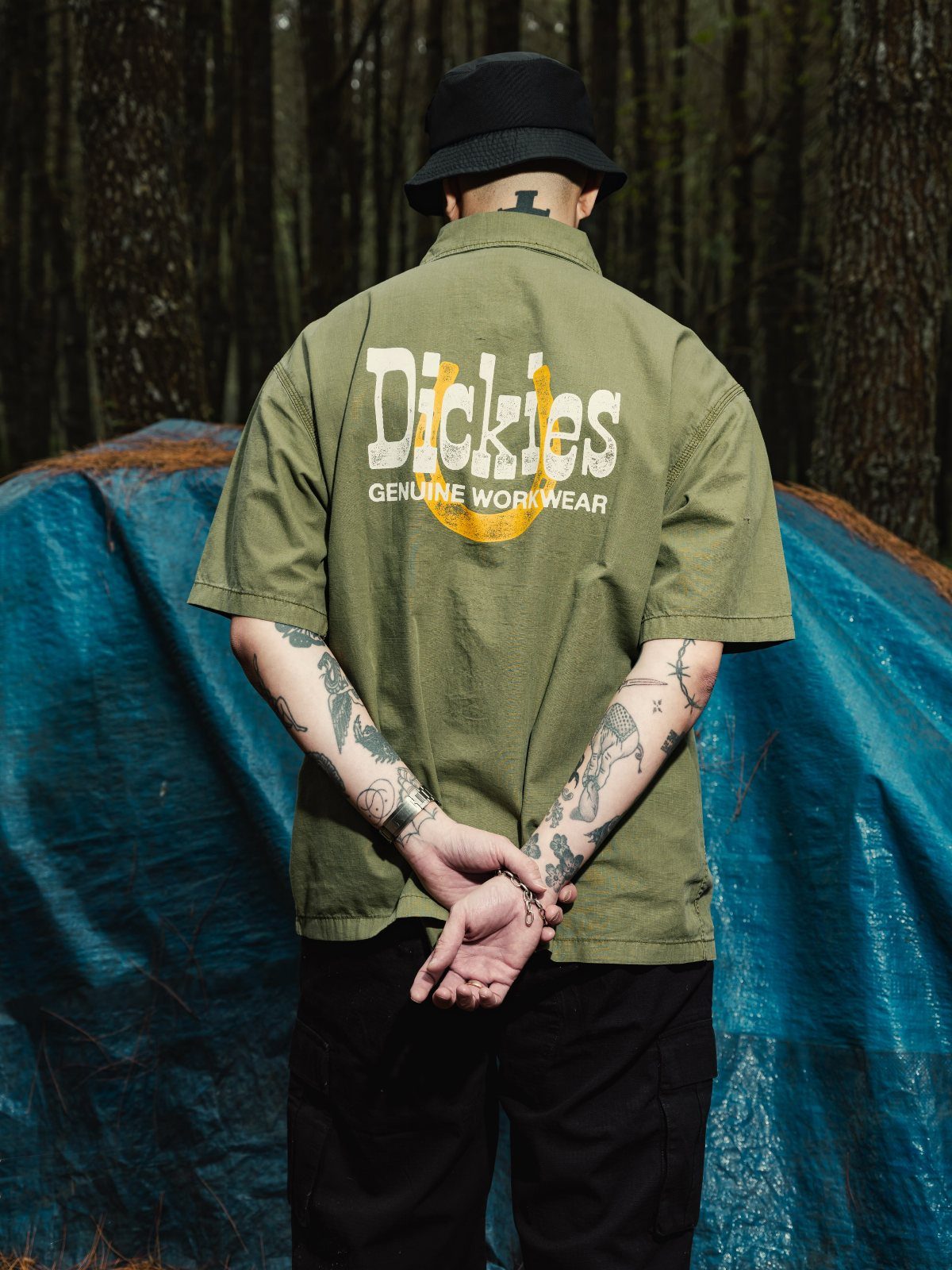 A person wearing an olive green Dickies work shirt and black bucket hat standing in a forest.
