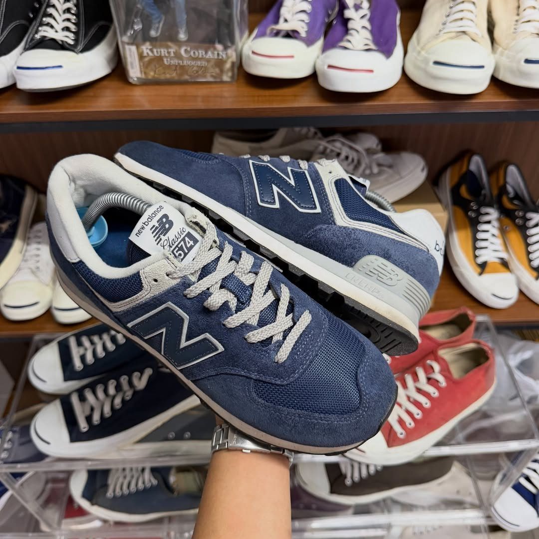 A hand holding up a pair of navy blue New Balance 574 sneakers in a shoe store.