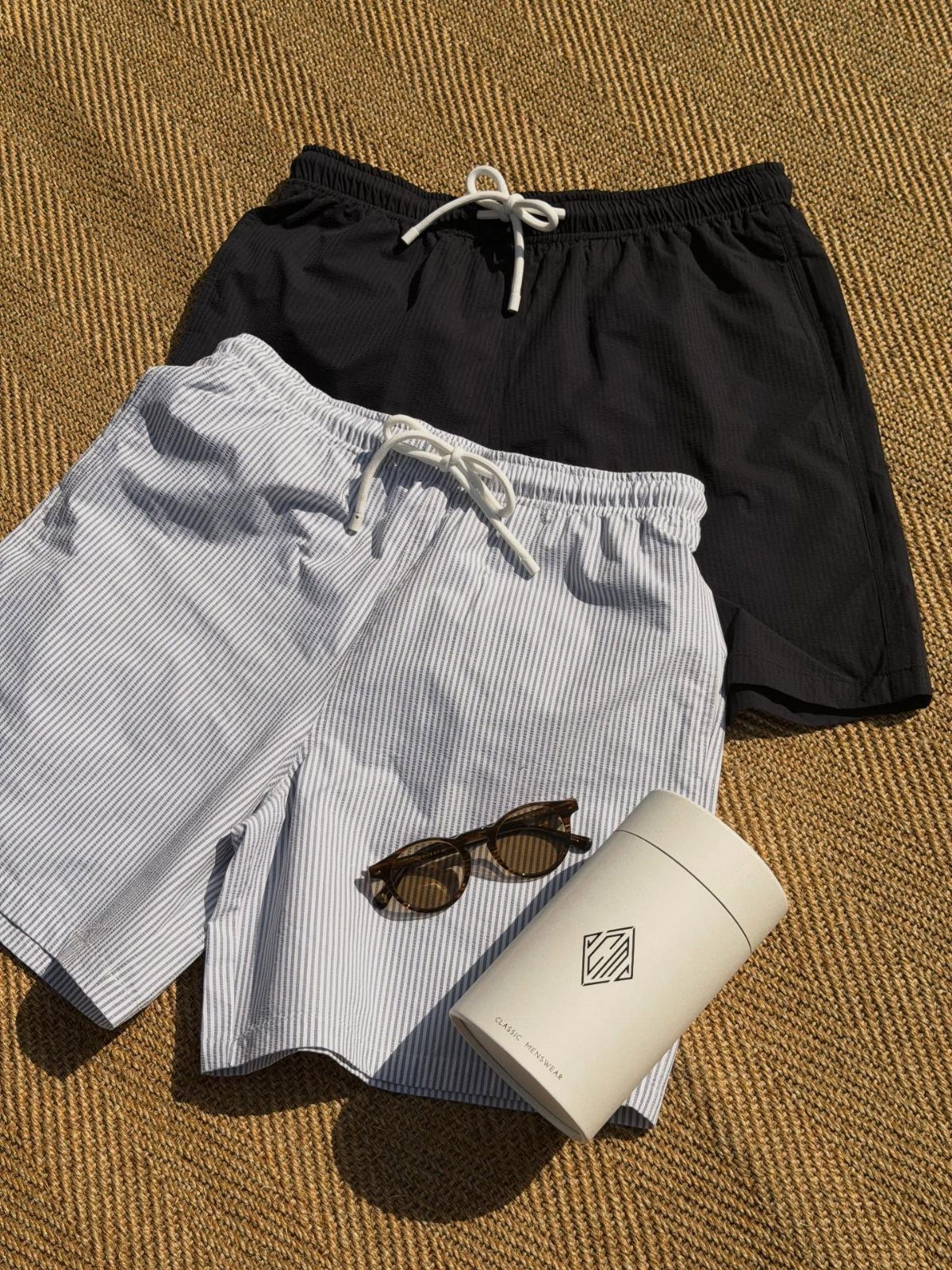 Light blue striped seersucker shorts and black shorts with sunglasses on a tan textured surface.