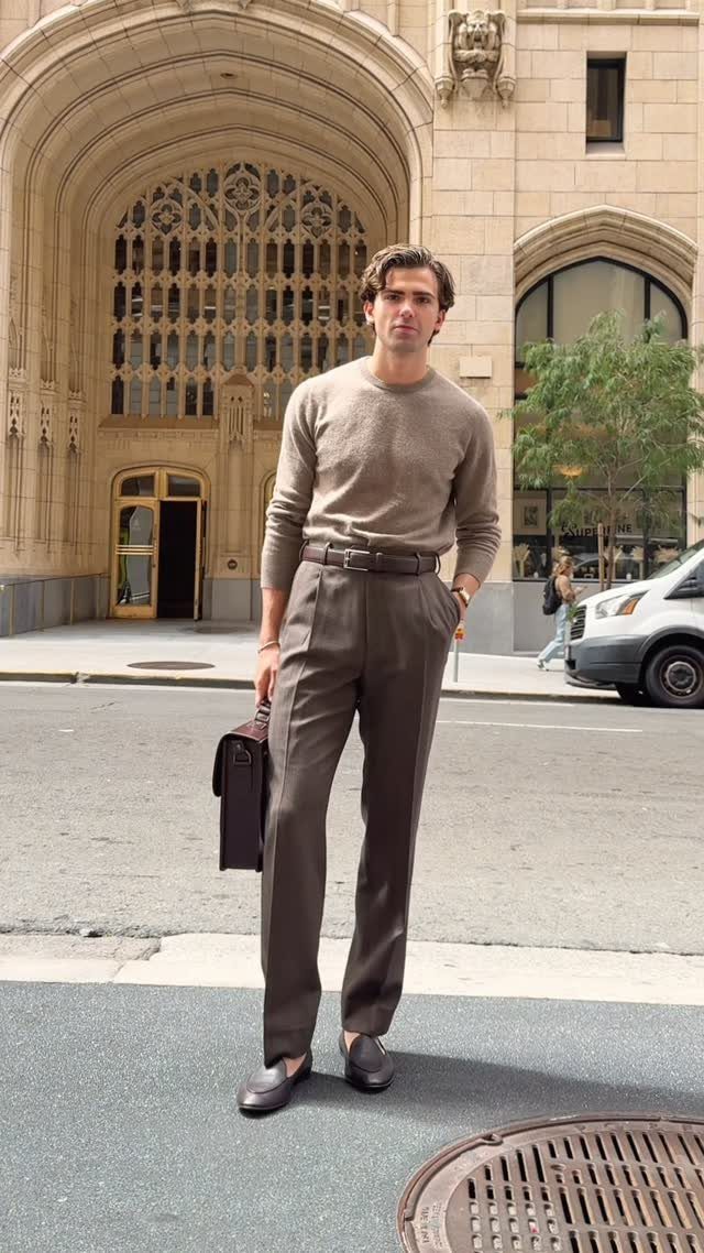 Man in a beige sweater and brown trousers standing on a city street holding a briefcase.