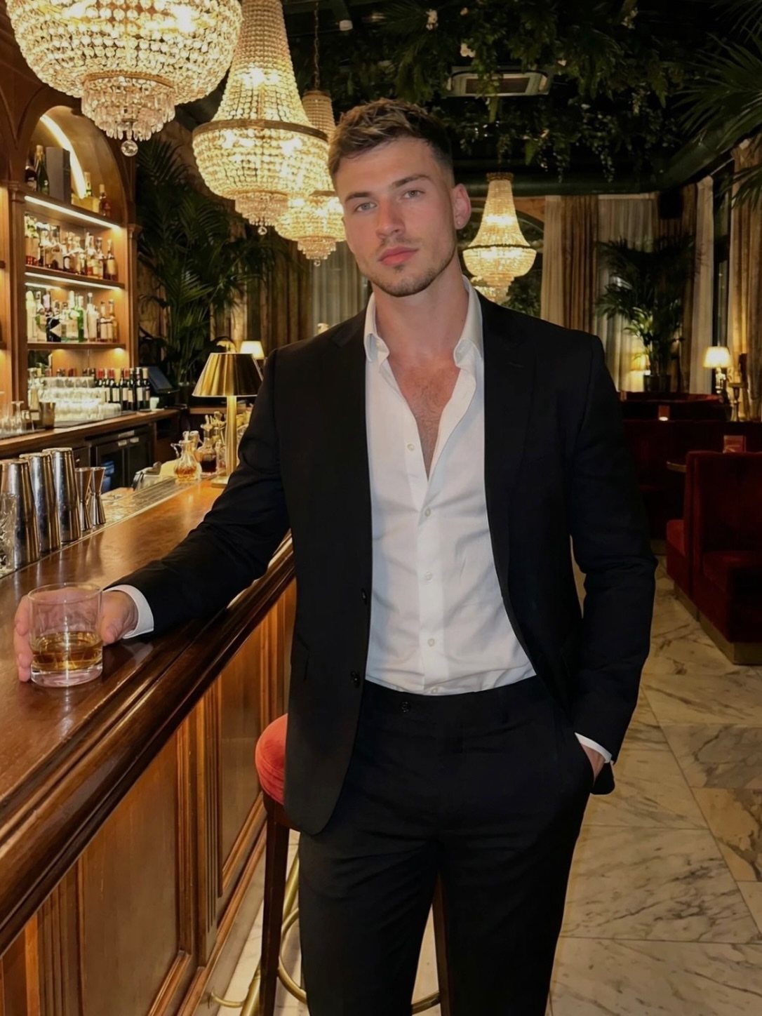 A man wearing a black suit and white shirt stands at a bar holding a drink.