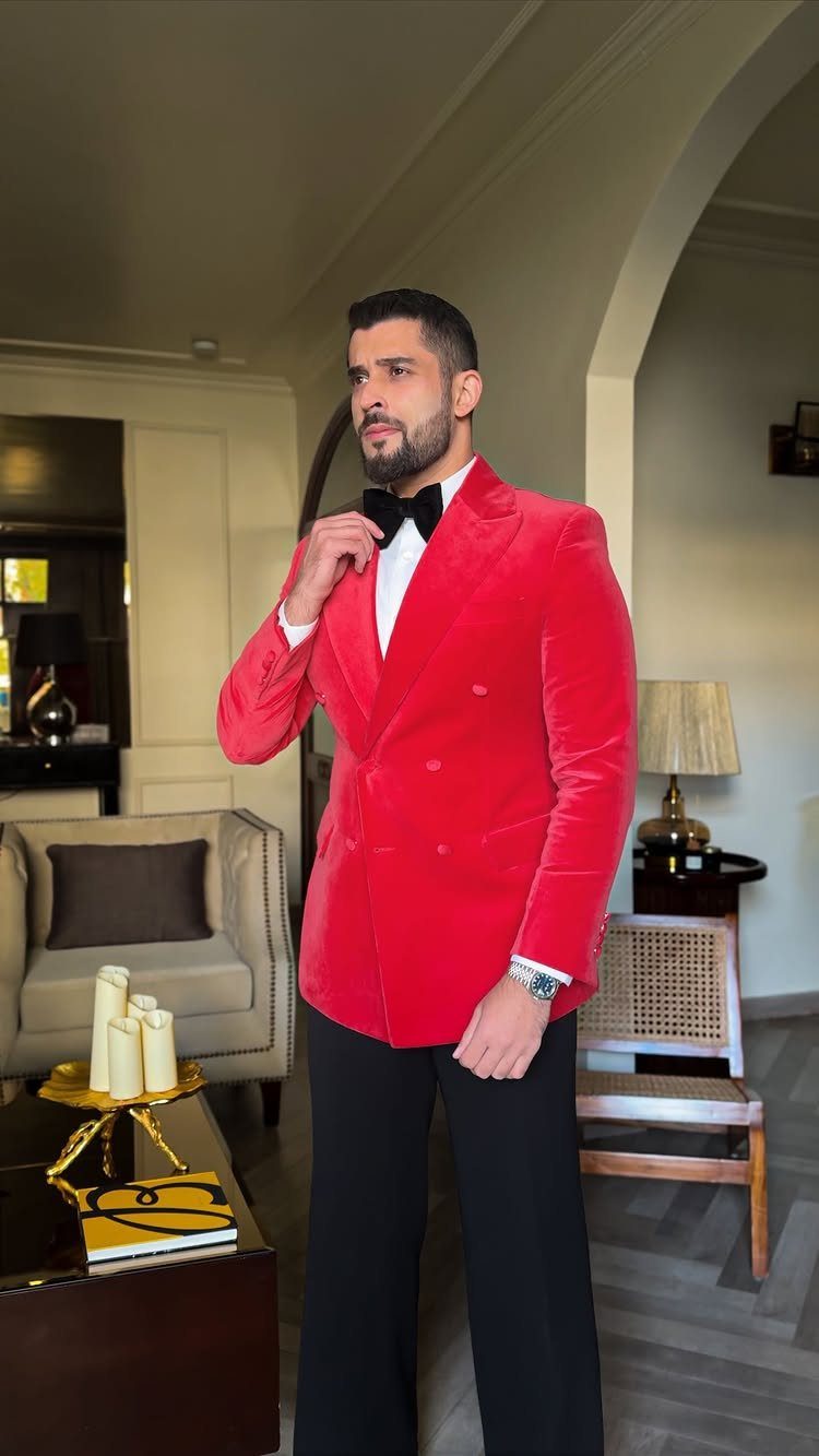 Man wearing a red velvet blazer, white shirt, black bow tie, and black trousers standing indoors.