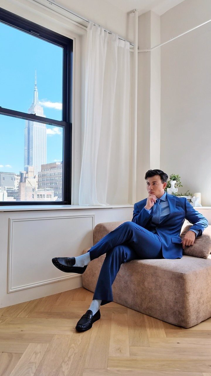 Man in a blue suit sitting on a tan sofa near a large window overlooking New York City.