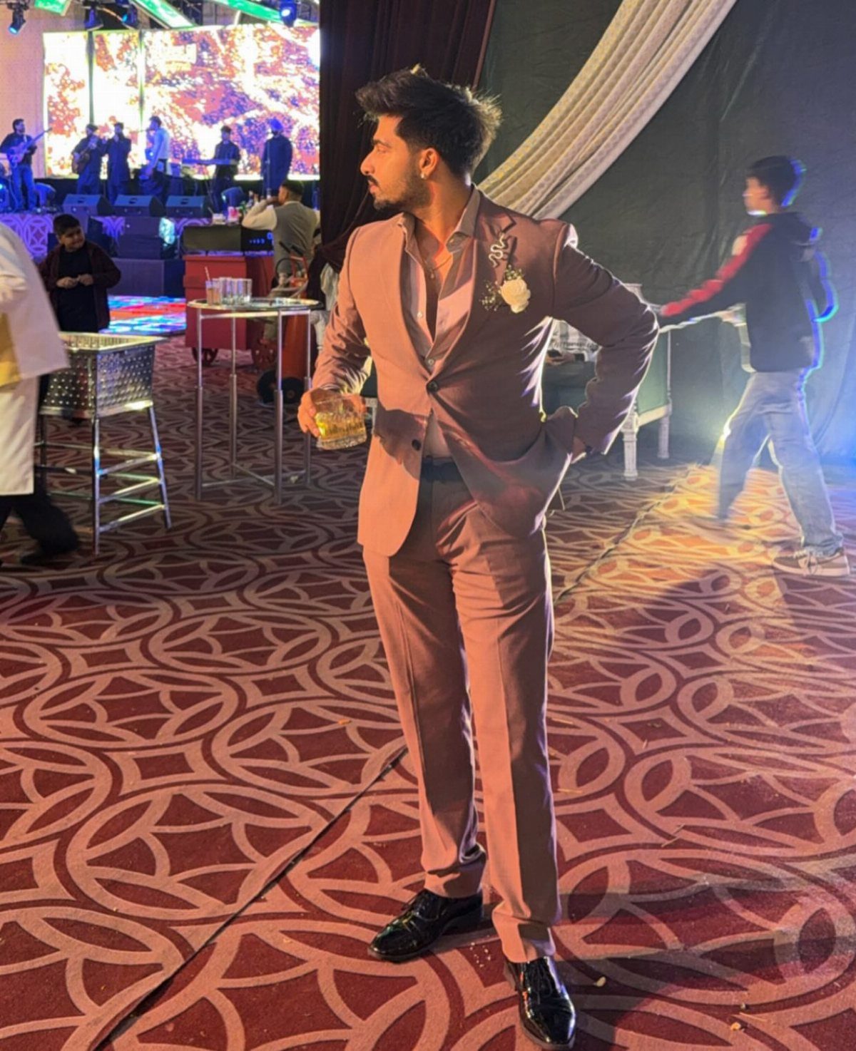 Man in a mauve tailored suit posing in a decorated event hall with patterned red carpet.