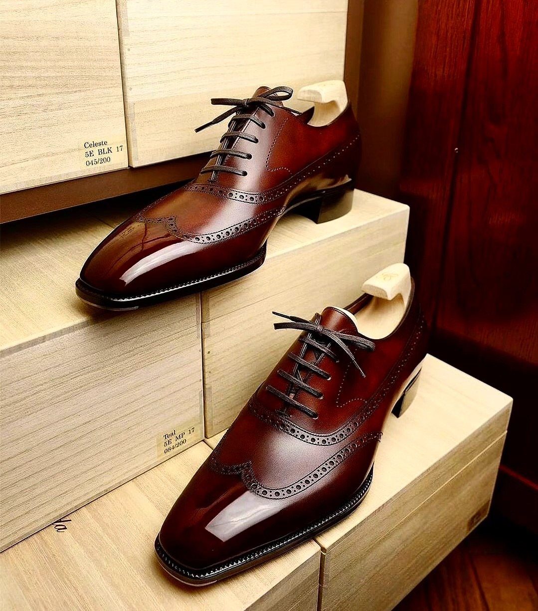 Polished burgundy leather wingtip derby shoes resting on light wooden shoeboxes.