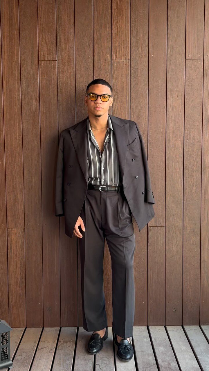 Man in a black suit and striped shirt posing against a dark wooden wall.