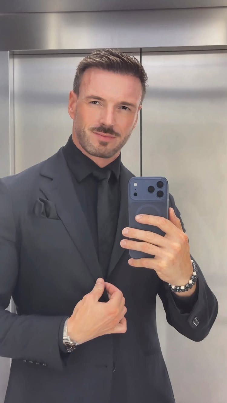 Man in a dark charcoal suit and black tie taking a mirror selfie.