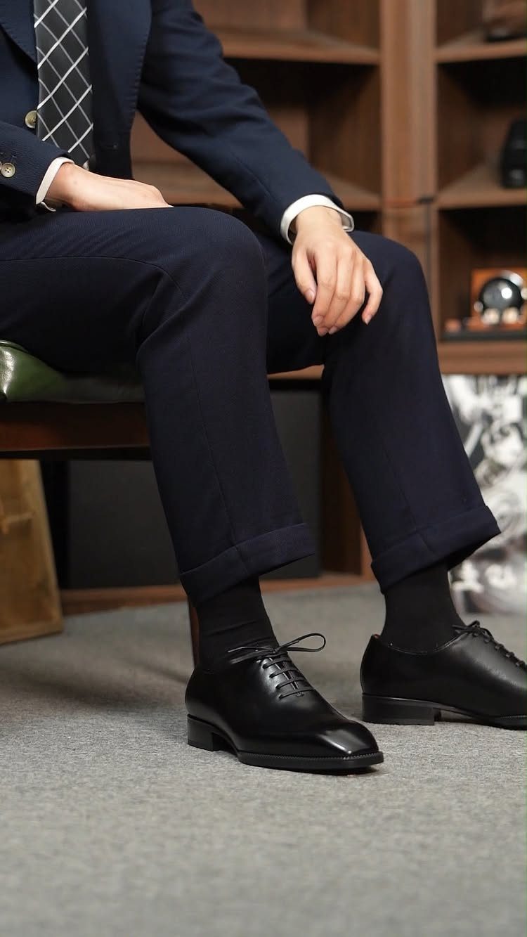 Man wearing navy trousers with cuffed hems, black socks, and black leather derby shoes.
