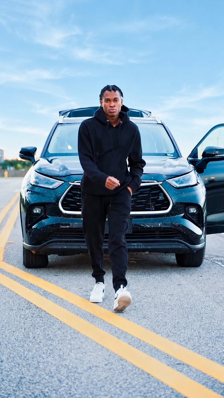 Man in black hoodie and joggers walking on a road in front of a dark SUV.