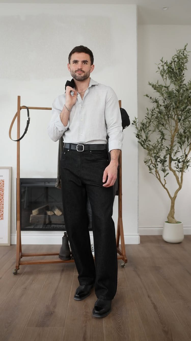 Man posing in a light grey button-down shirt and black trousers indoors.