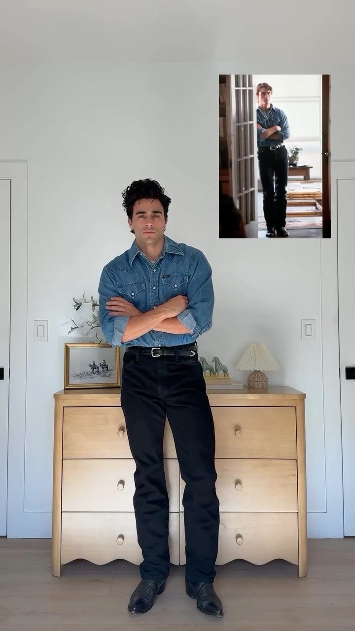 Man in a denim shirt and black trousers posing in a bright room.