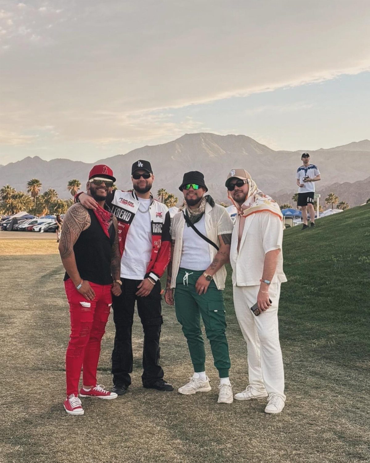 Four men posing in various streetwear outfits in a grassy field with mountains behind them.