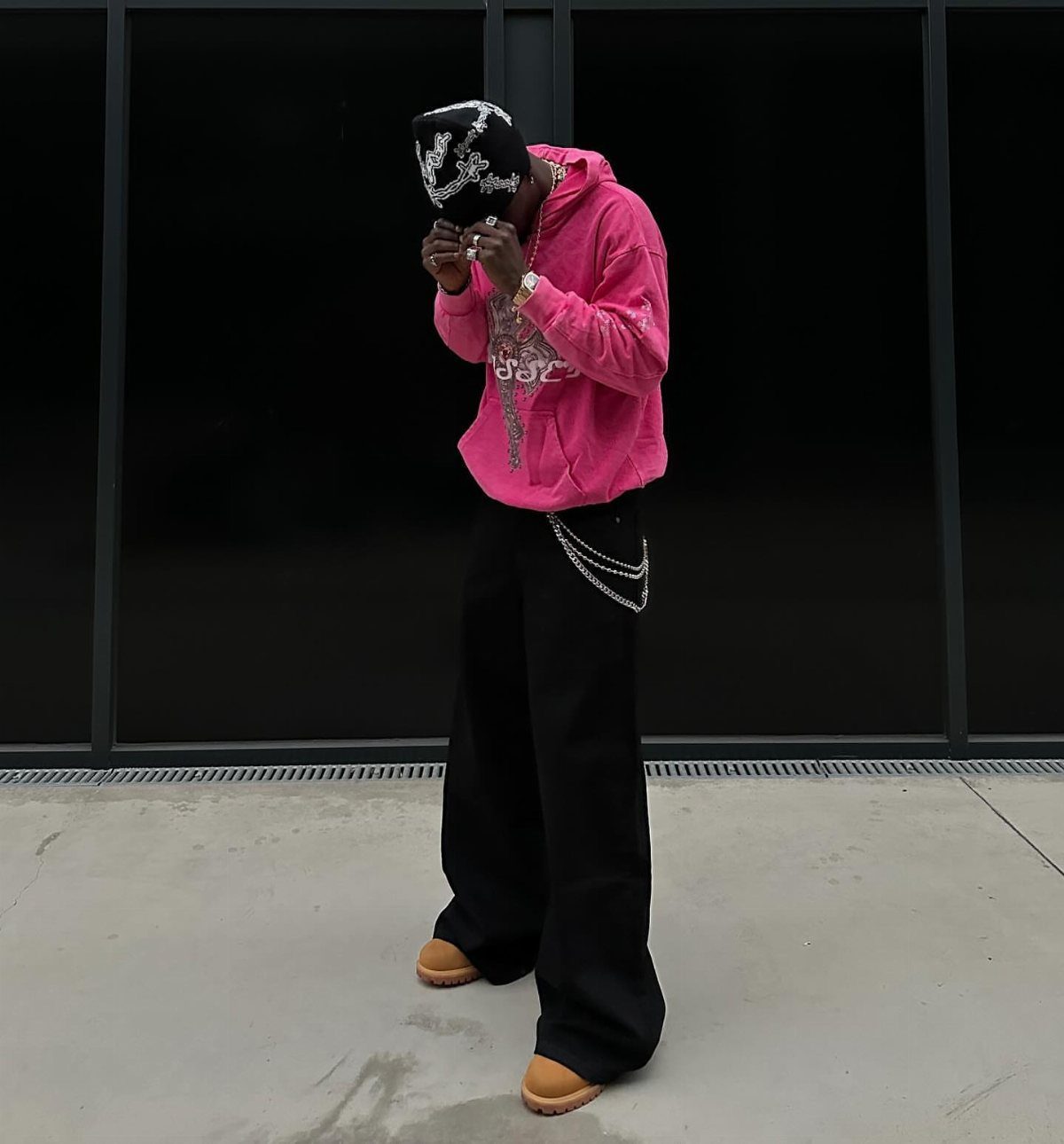 A person in a pink graphic hoodie and black wide-leg trousers standing outdoors.