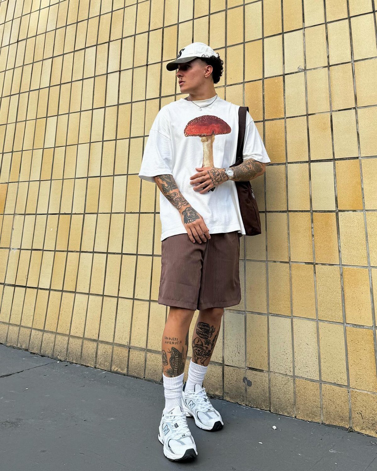 Man in white mushroom graphic t-shirt and brown shorts standing against a yellow tiled wall.