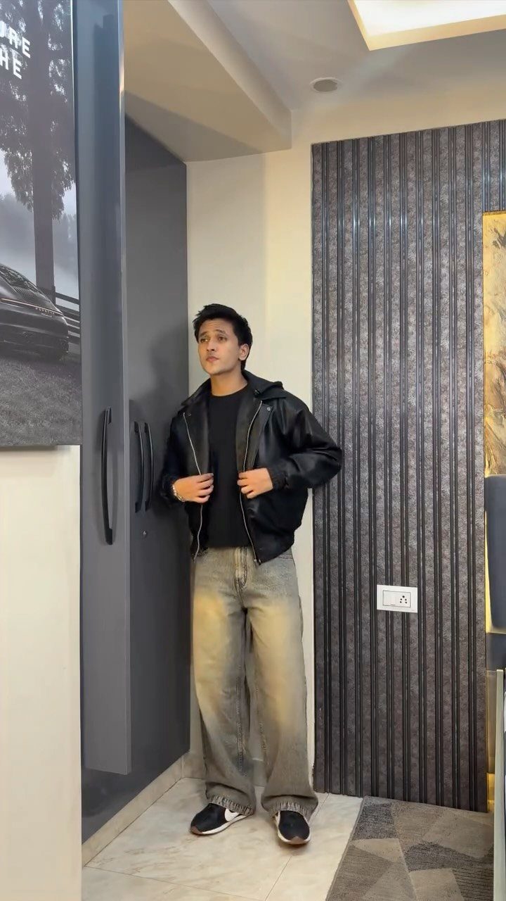 A young man posing in a black leather jacket, black t-shirt, and baggy grey jeans.