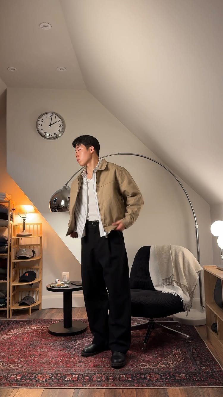 A man posing in a tan jacket, white shirt, and black trousers in a modern room.