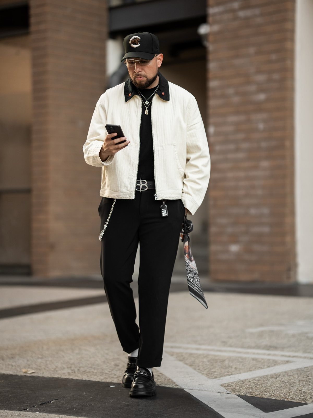 Man in a white corduroy jacket, black trousers, and a baseball cap walking while using a phone.