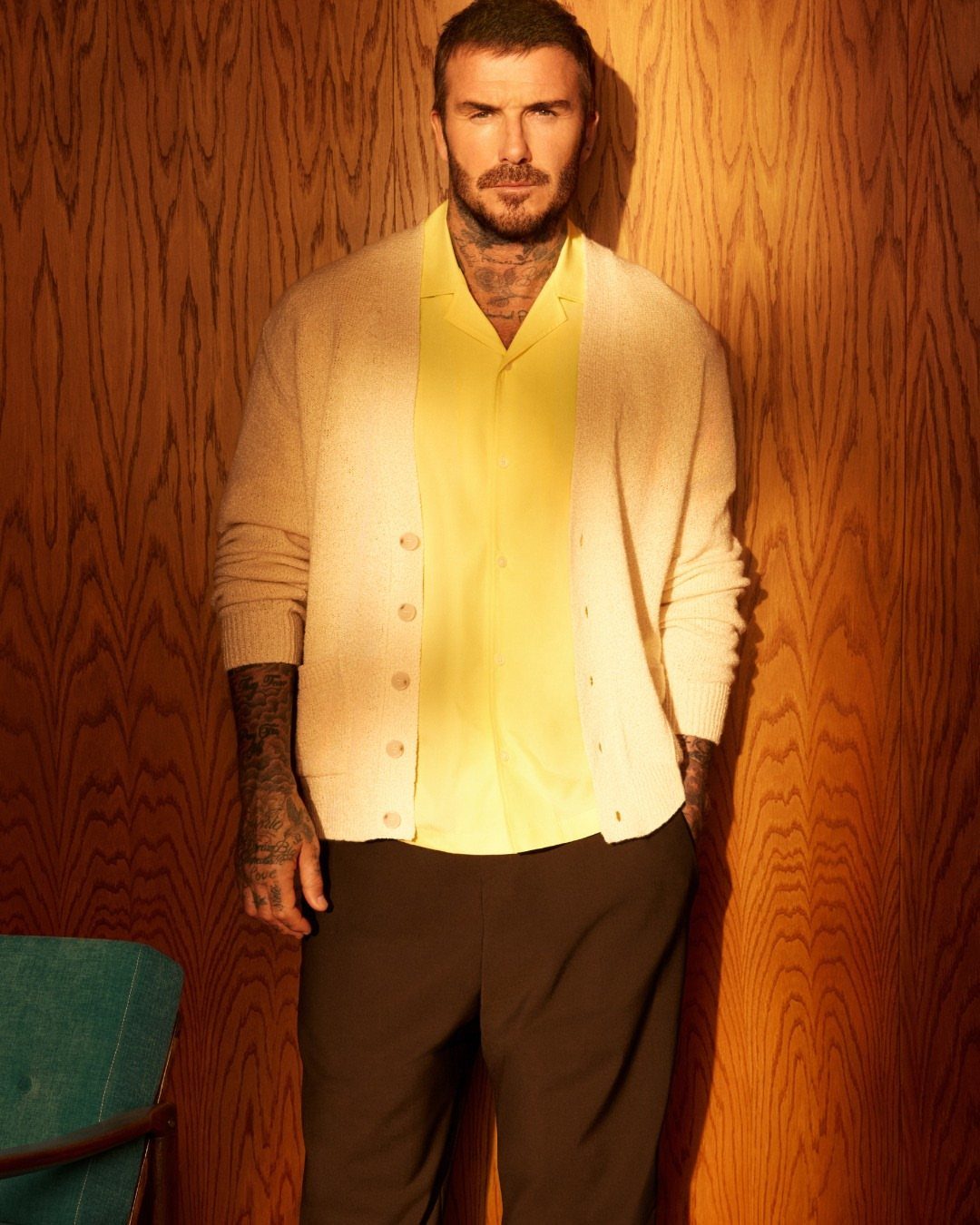 Man with tattoos wearing a yellow shirt and cream cardigan against a wood-paneled wall.